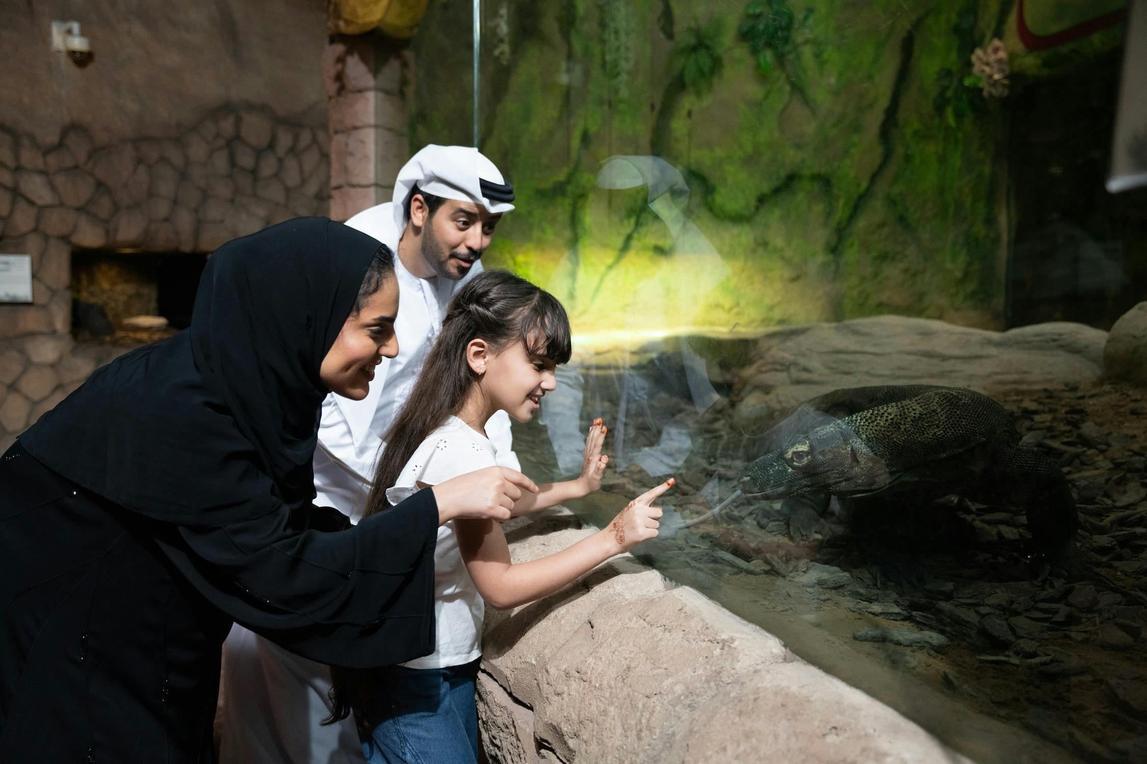 Three people, two adults and a child, look at an exhibit in an indoor environment with stone walls and greenery.