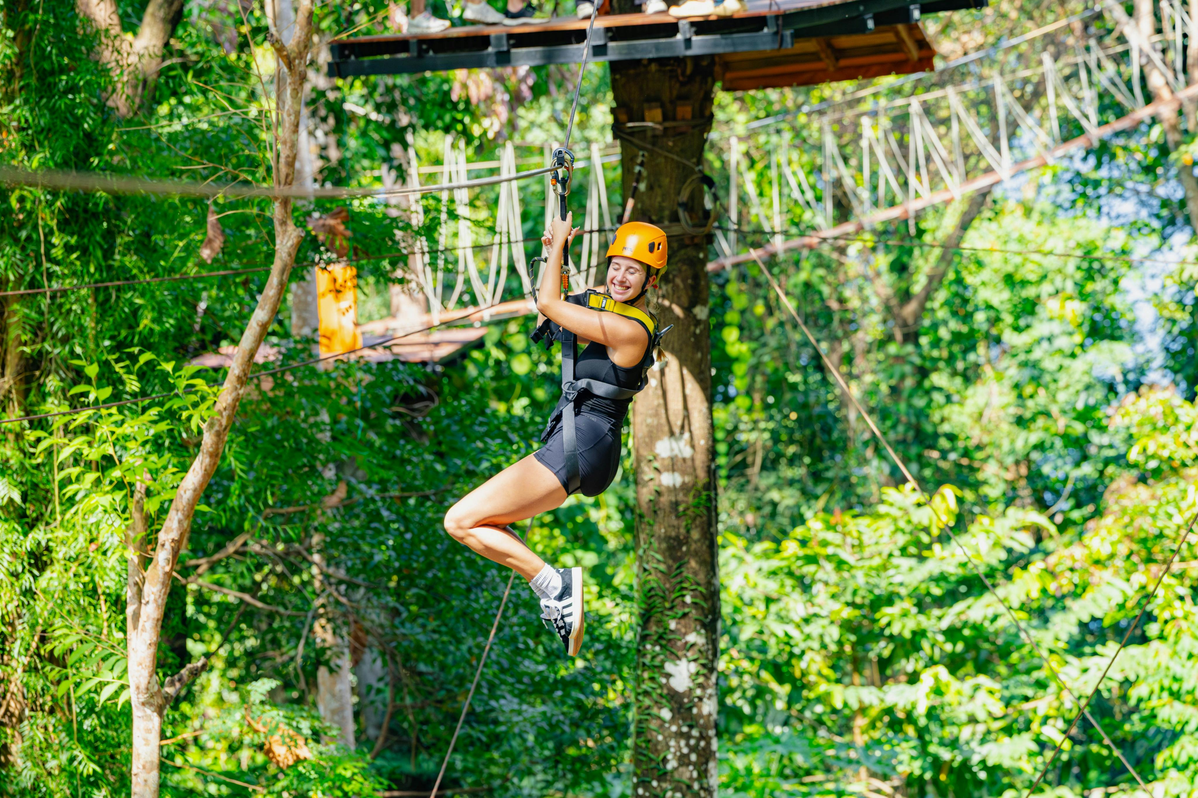 Person ziplining through a forested area, wearing a helmet and harness, smiling and mid-motion.