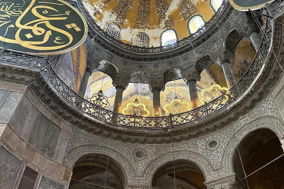 Interior of a historic building with ornate columns, a decorative dome, intricate carvings, and hanging chandeliers.