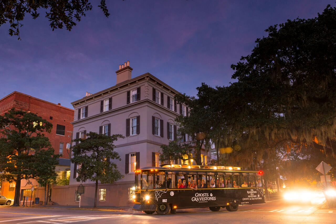A black tour trolley with "Ghosts & Gravestones" signage in front of a historic building at dusk, illuminated by streetlights.