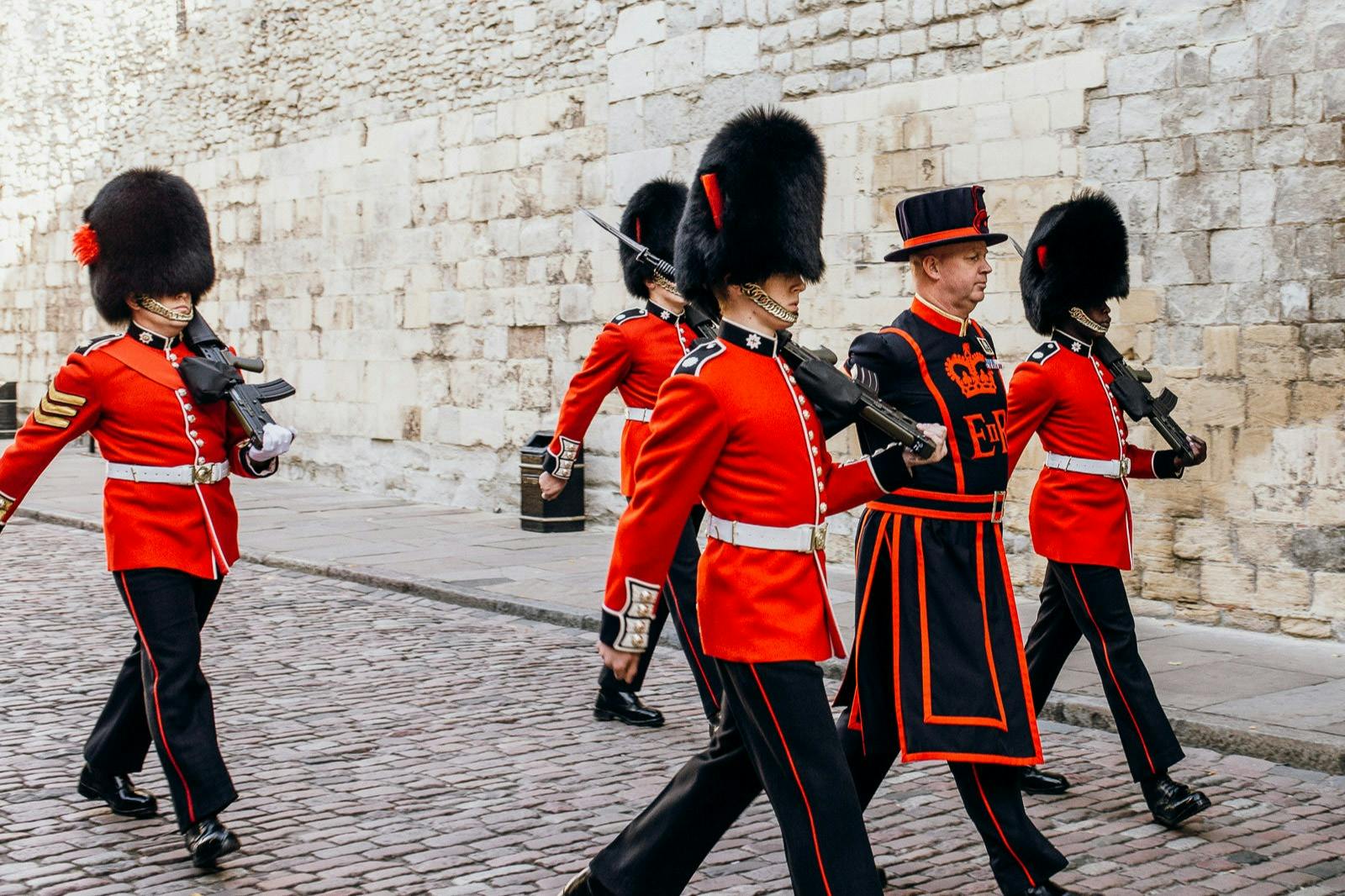 Three guards in red uniforms and bearskin hats march alongside a man in ceremonial dress by a stone wall.