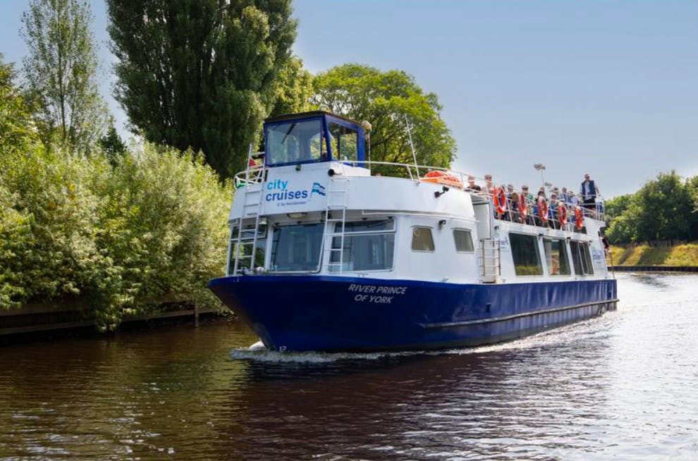 Een rondvaartboot met de naam "River Prince of York" met veel passagiers vaart op een rivier, omringd door bomen.