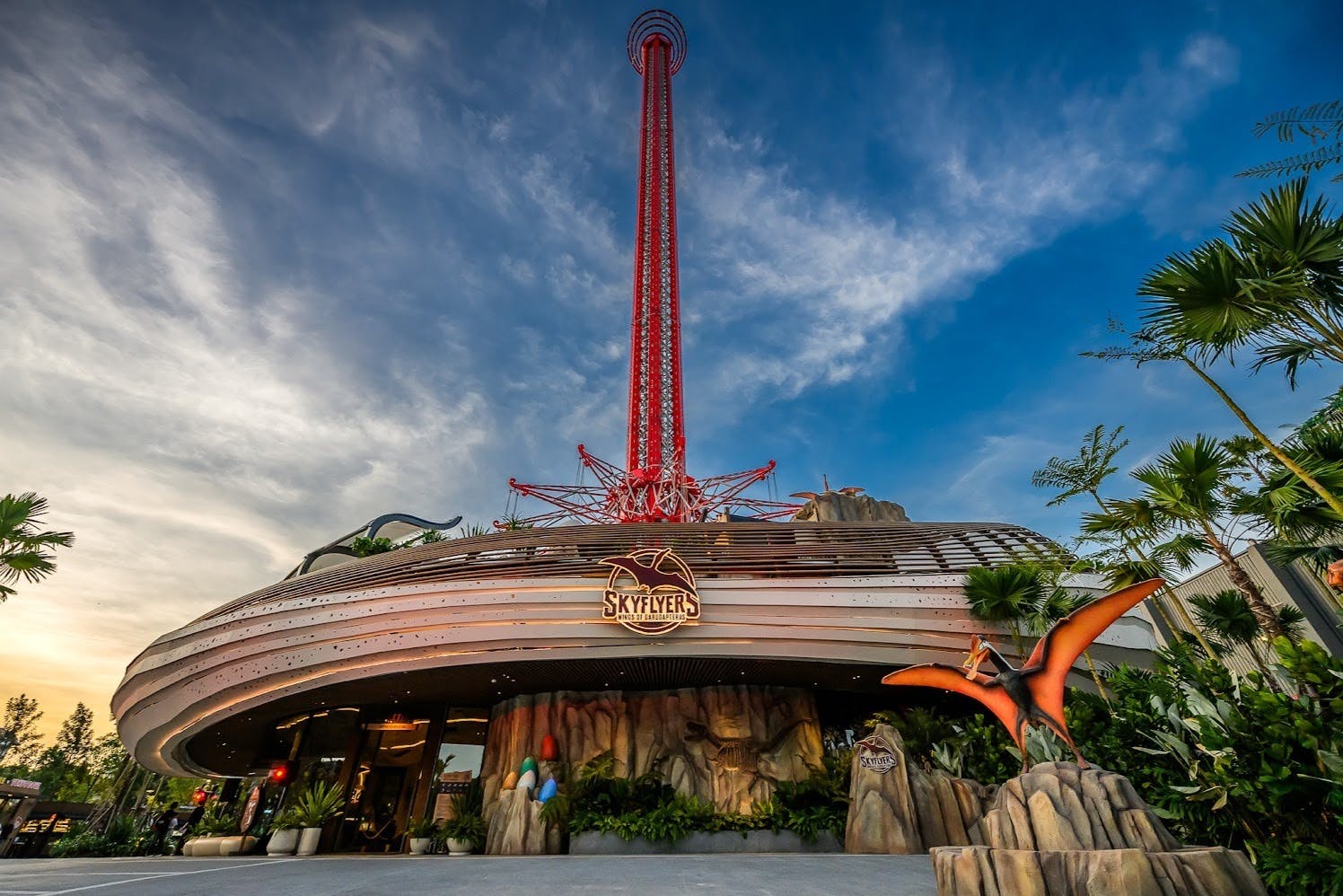 A tall red amusement ride named Skyflyers, with a rocky-themed entrance and a pterosaur statue in front under a blue sky.