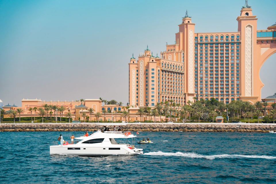 A luxury resort with palm trees beside a body of water, with a white yacht and a jet ski on the water in the foreground.