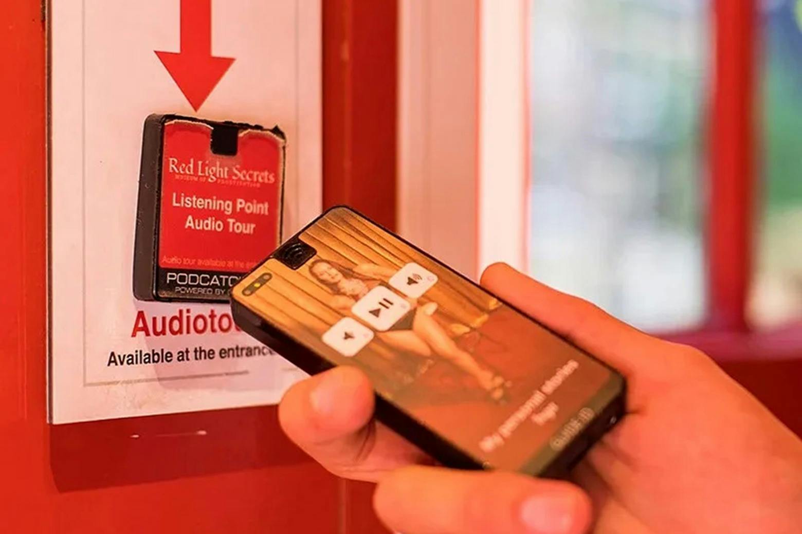 Mano che tiene uno smartphone che mostra un'app per il tour audio davanti al cartello del punto d'ascolto del museo "Red Light Secrets".