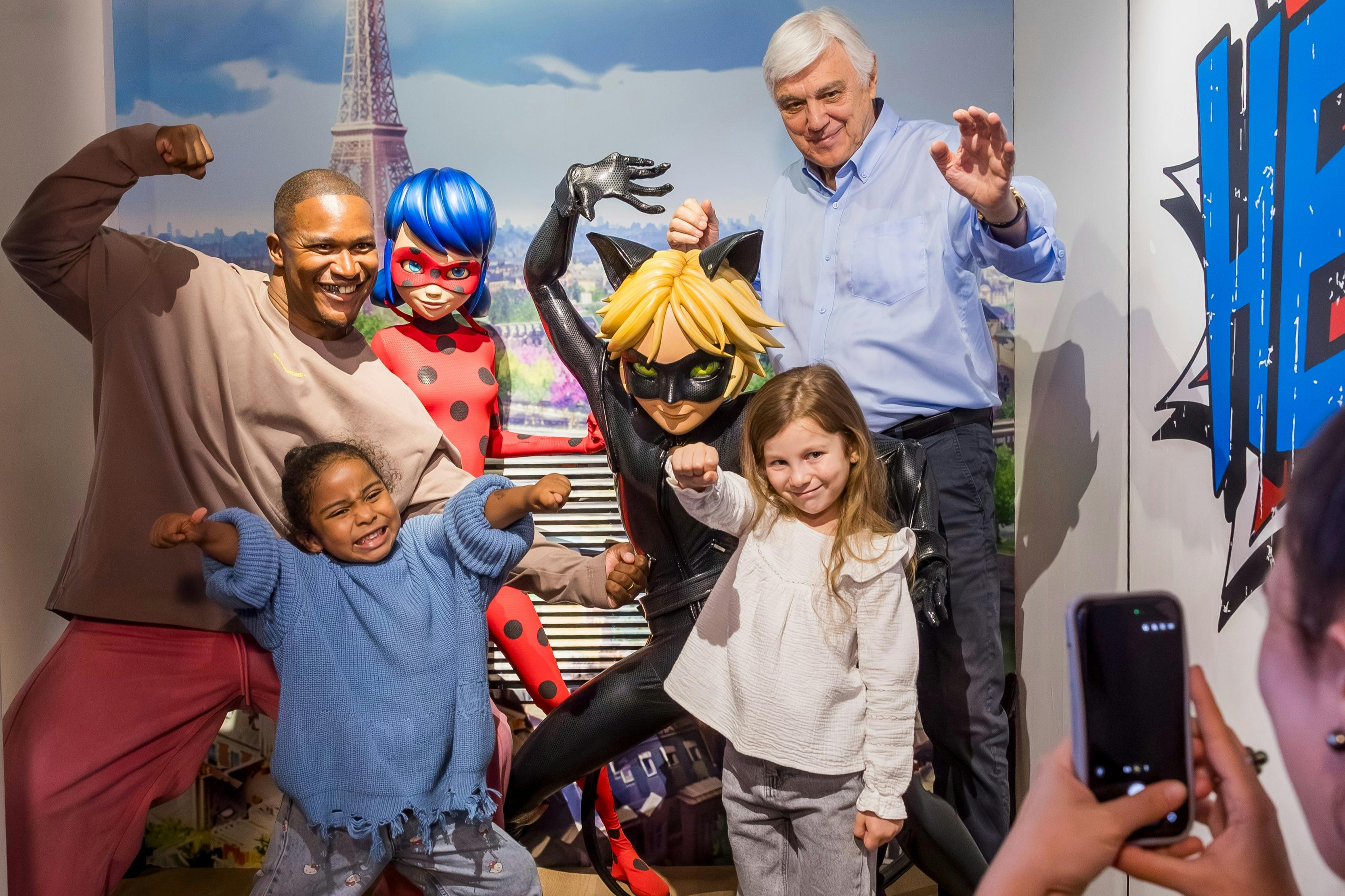 Zwei Erwachsene und zwei Kinder posieren mit kostümierten Figuren in einer Kulisse im Pariser Stil, darunter Ladybug und Cat Noir.