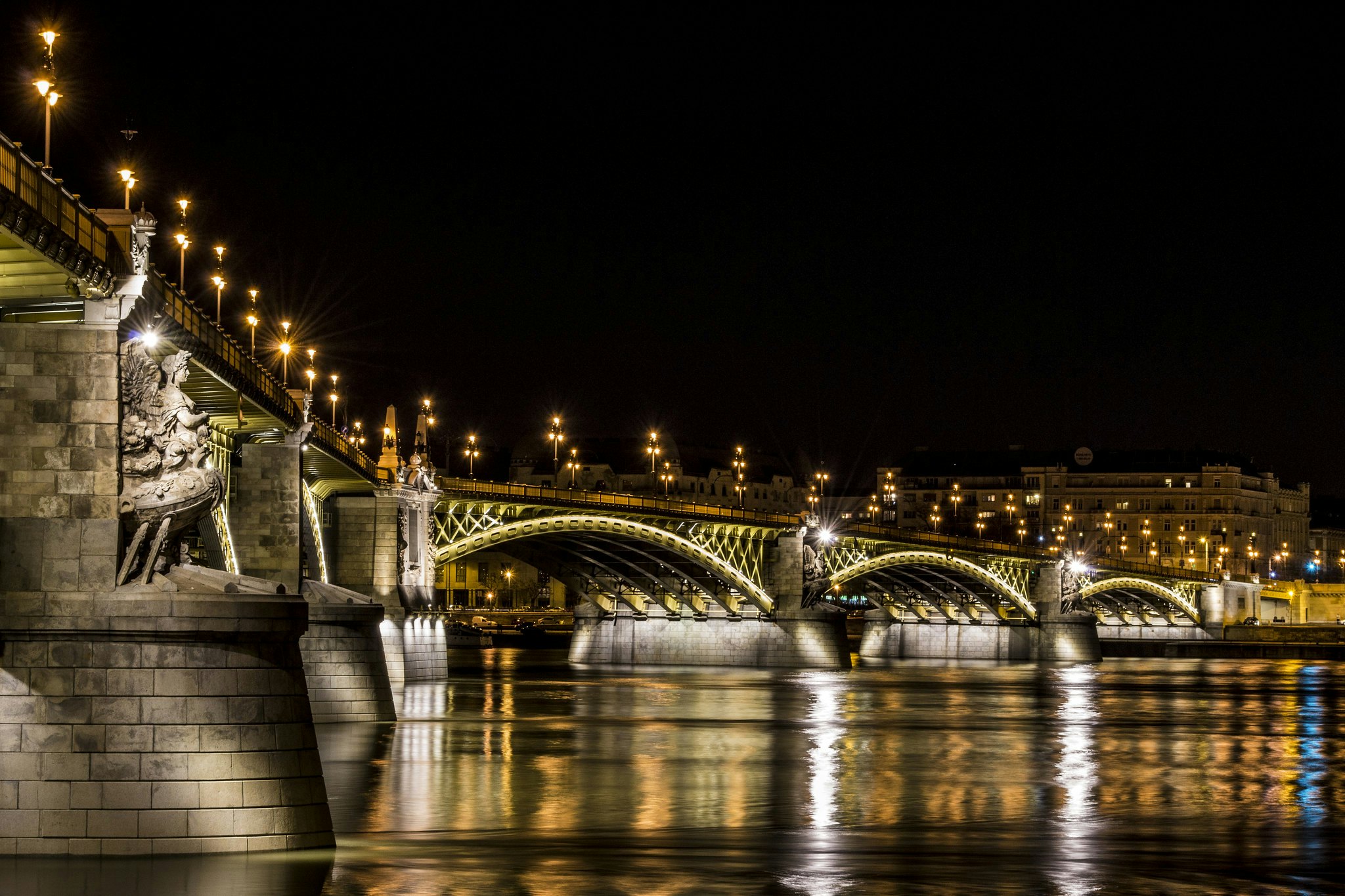 Ponte iluminada com esculturas ornamentadas, atravessando um rio à noite. Os edifícios e as luzes da cidade são visíveis em segundo plano.