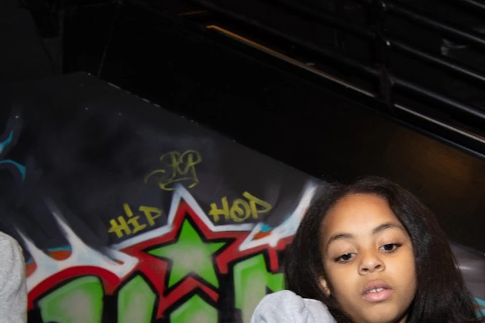 Two children and an adult at a DJ setup with turntables, graffiti background, and a Supreme sticker on the equipment.