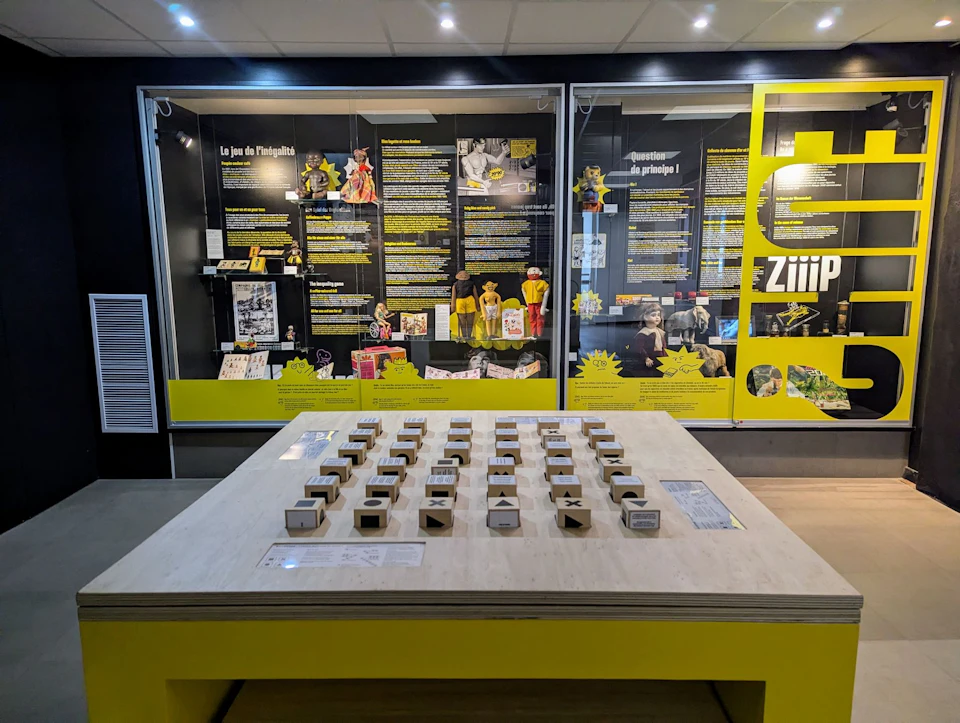 An exhibition featuring text panels, yellow accents and interactive game pieces on a table, all housed within a display room.