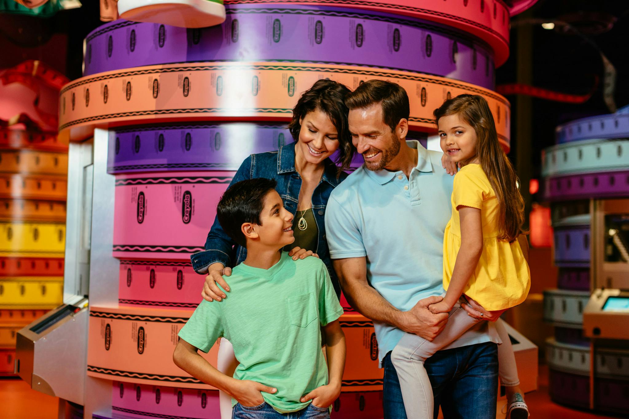 A smiling family of four stands together in front of large, colorful stacks of packing tape rolls.