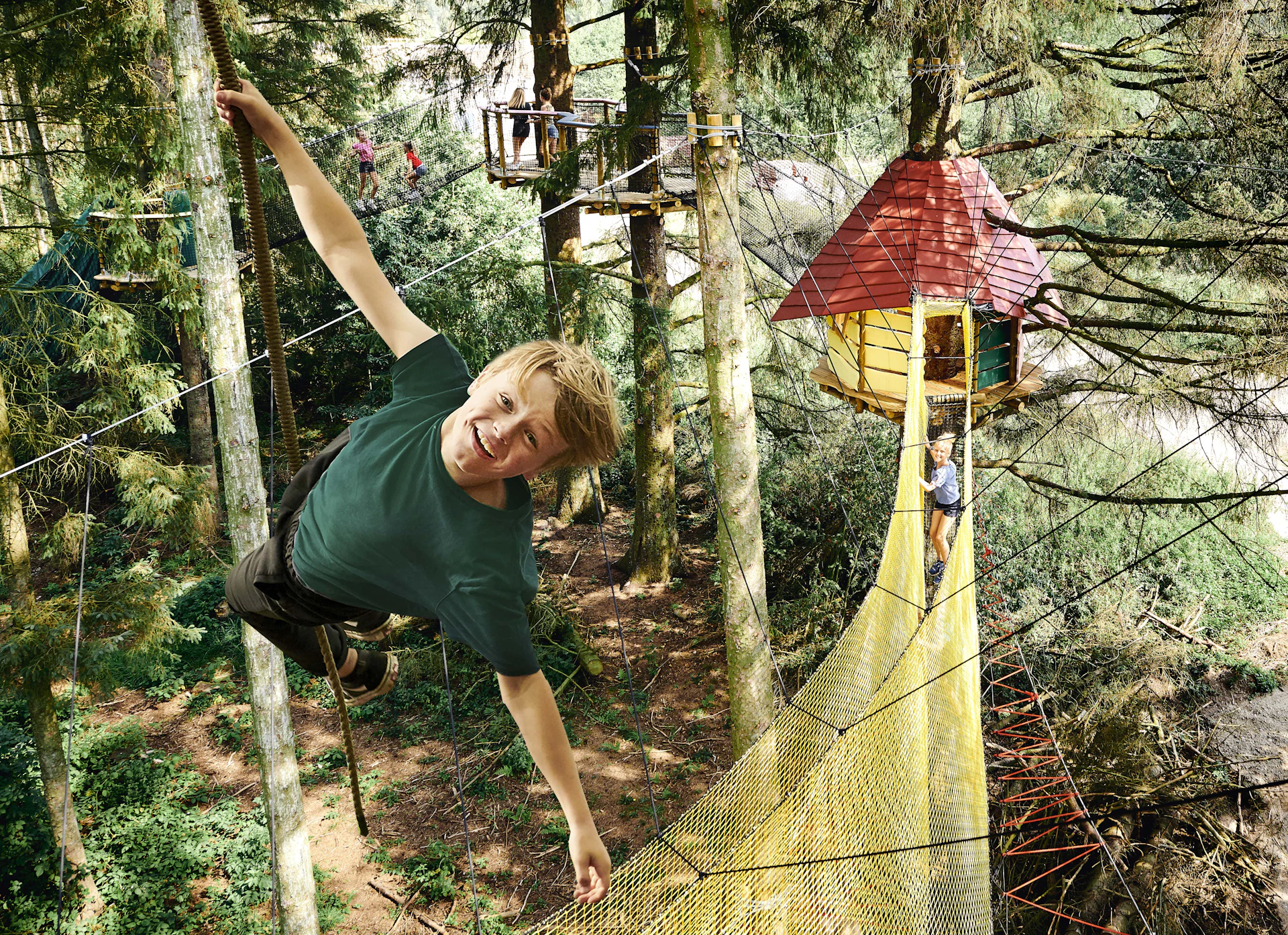 A smiling boy hangs onto a rope in a forest adventure park. A yellow net bridge leads to a treehouse in the background.