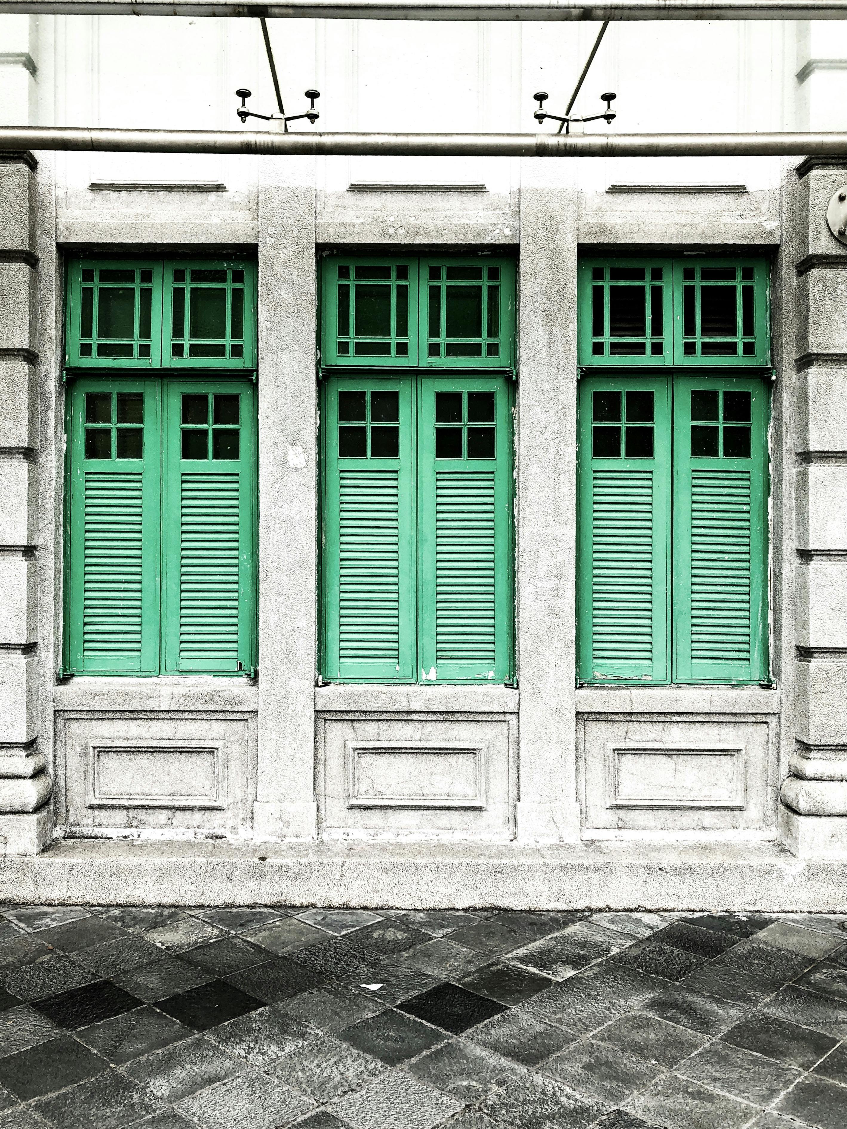 Facciata con tre finestre verdi con persiane incastonate in un muro di pietra grigia, con cornici rettangolari decorative sotto ogni finestra.