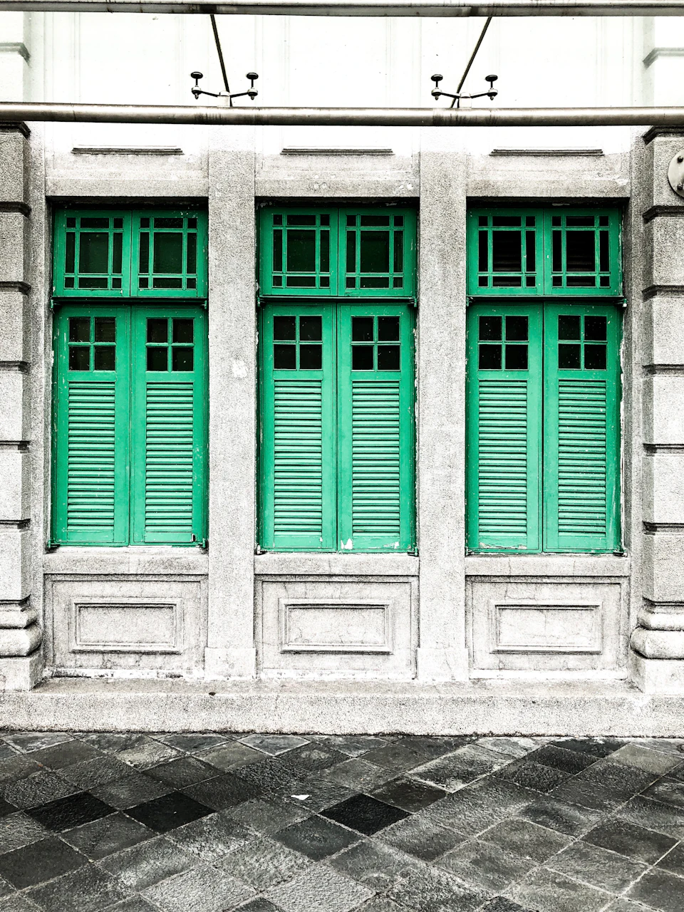Facade with three green, shuttered windows set in a gray stone wall, featuring decorative rectangular frames beneath each window.