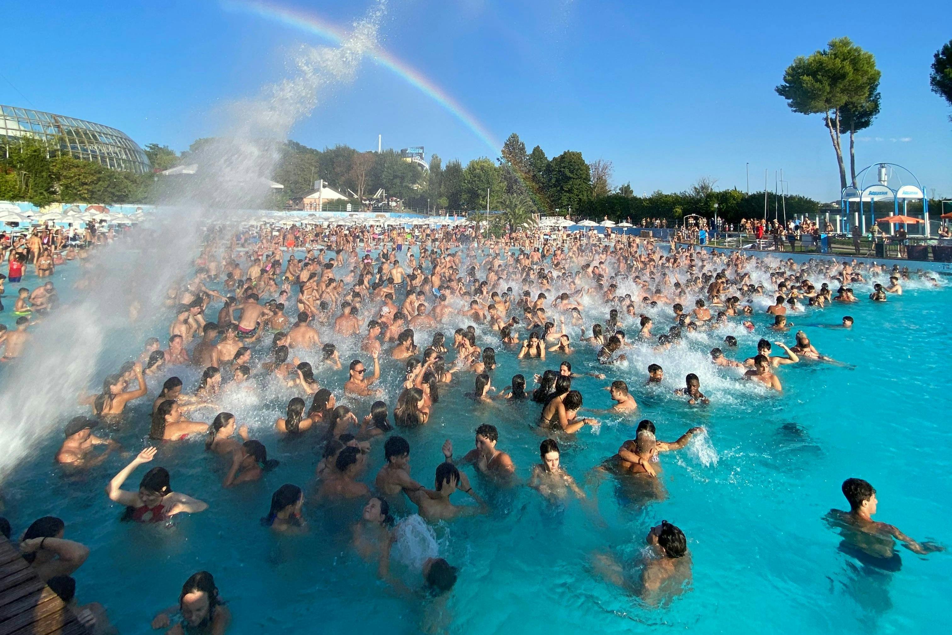 Una grande folla di persone che si diverte in una piscina a onde sotto un cielo azzurro e limpido con un arcobaleno visibile sullo sfondo.