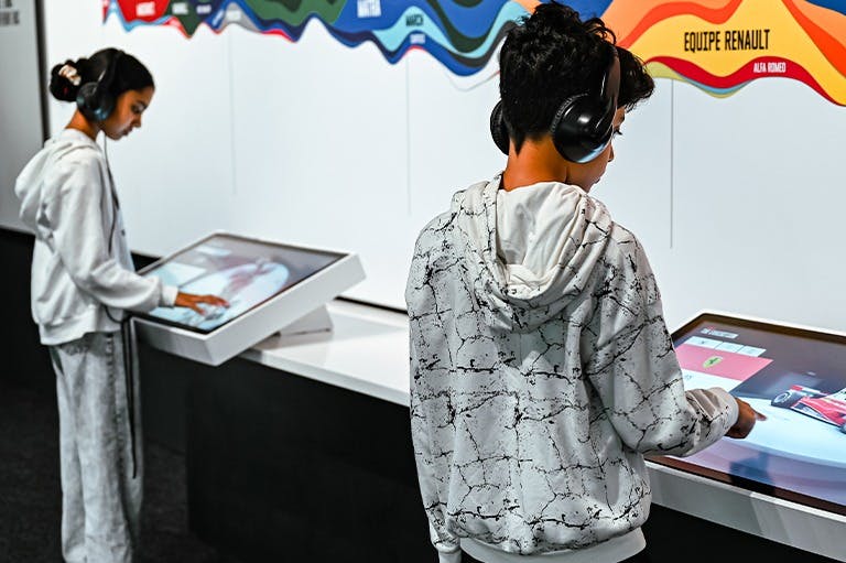 Dos nens amb auriculars interactuen amb pantalles tàctils en una zona d'exposició moderna i ben il·luminada.