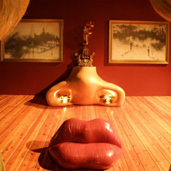 Surrealistiskt rum med en stor, läppformad soffa, en nässkulptur och inramade målningar på en röd vägg.