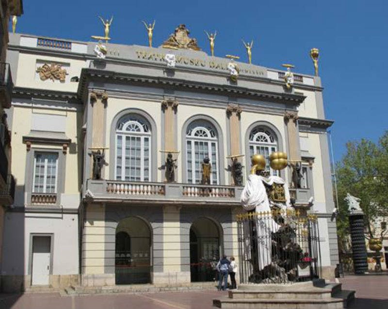 Ornate building facade with large arched windows, statues, and gold accents, fronted by a sculptural fountain. Clear blue sky above.