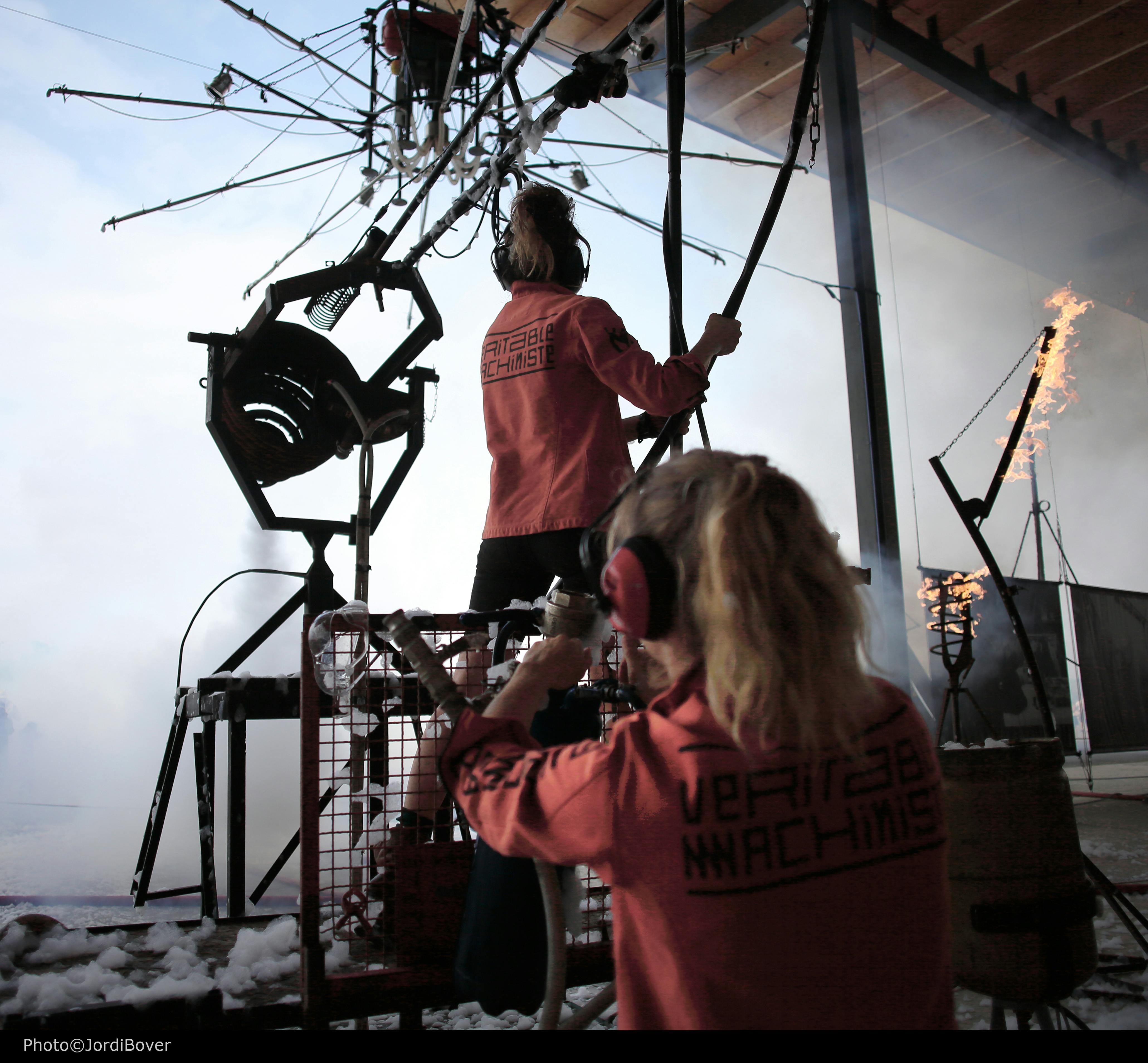两个穿着匹配红色夹克的人操作一个复杂的机械设置，带有各种杠杆和燃烧的火焰。