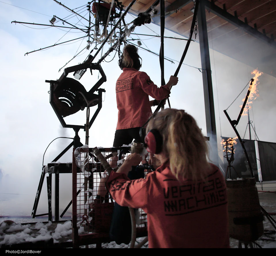 Two people in matching red jackets operate a complex, mechanical setup with various levers and burning flames.