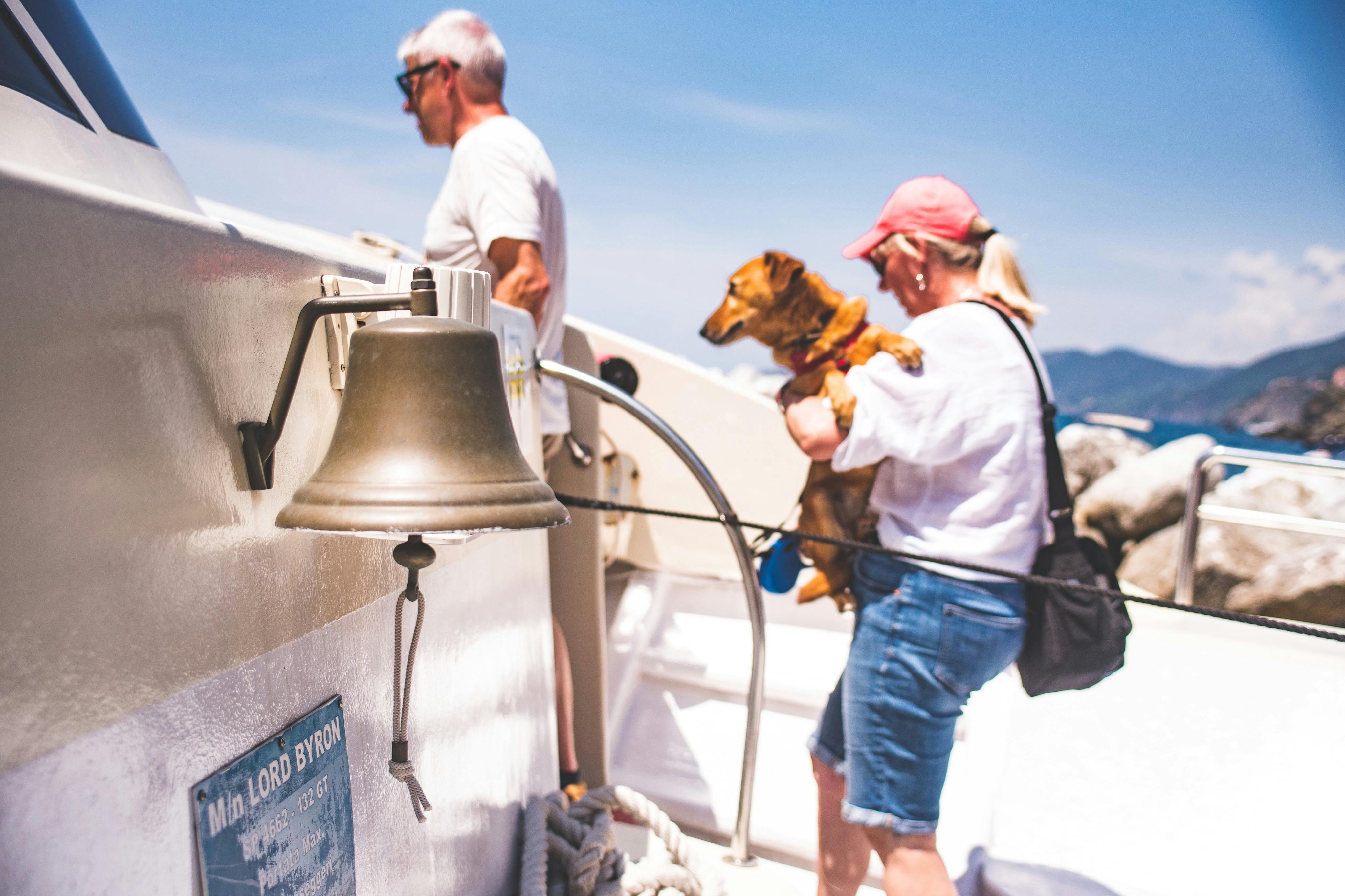 Δύο άνθρωποι σε μια βάρκα, ο ένας κρατώντας έναν σκύλο, με ένα ορατό κουδούνι στο προσκήνιο κάτω από έναν καταγάλανο ουρανό.