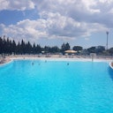 Grande piscina all'aperto con pochi bagnanti, circondata da alberi sotto un cielo parzialmente nuvoloso.