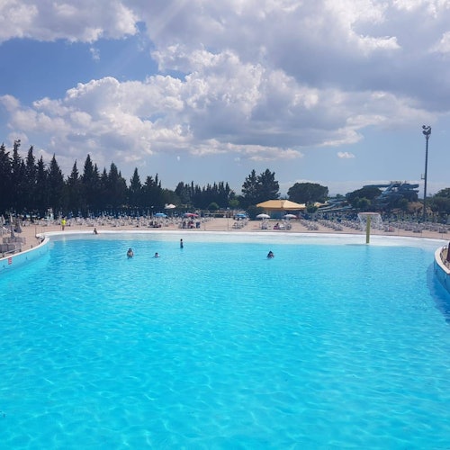 Grande piscina all'aperto con pochi bagnanti, circondata da alberi sotto un cielo parzialmente nuvoloso.