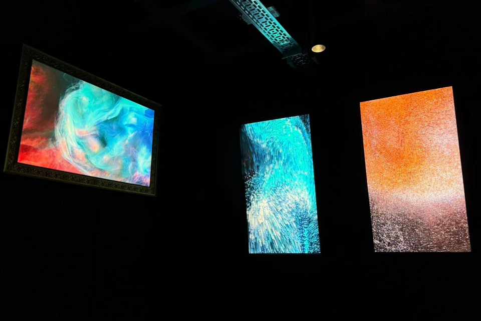 Three illuminated abstract artworks in a dark room, each depicting colorful and textured patterns in blue, orange, and multicolor.