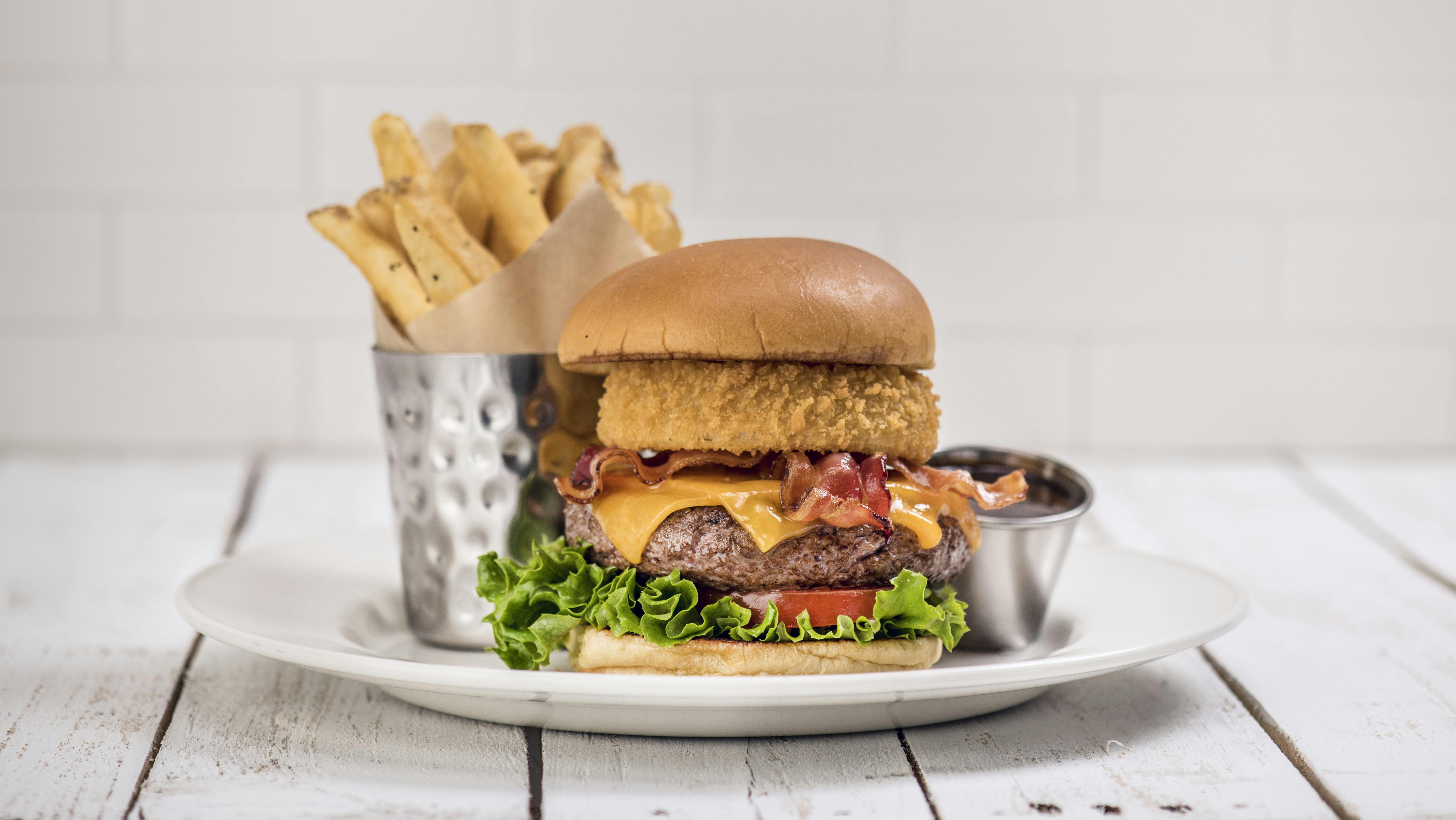 Burger se slaninou, sýrem, cibulovým kroužkem, salátem a rajčetem na bílém talíři s hranolky v kovovém šálku a malou miskou omáčky.