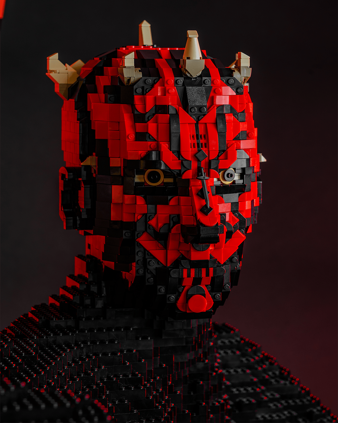 Scultura LEGO di una figura con il volto rosso e nero, occhi gialli e piccole corna, su uno sfondo scuro.