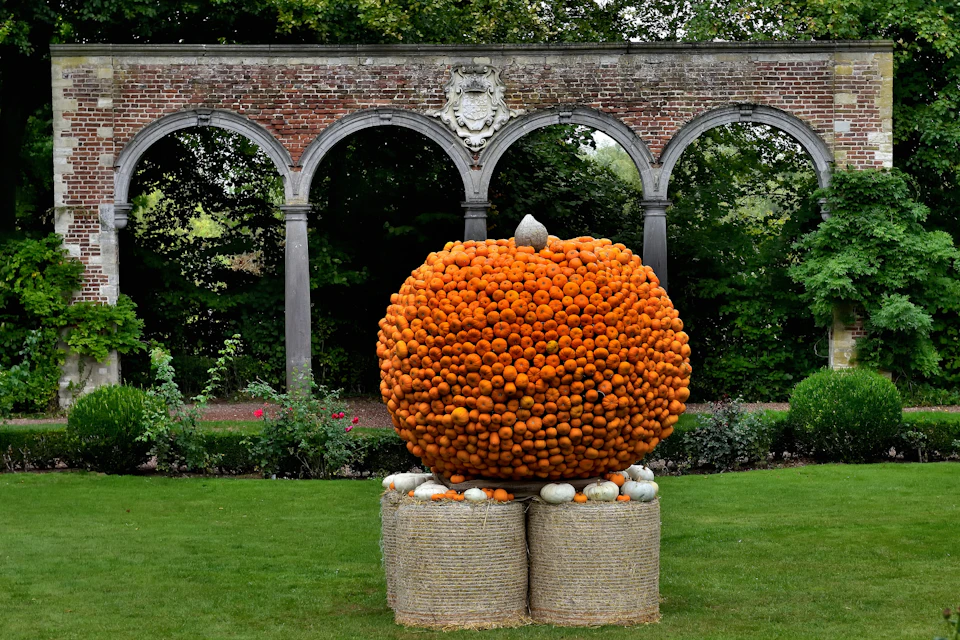 A large pumpkin sculpture made of small pumpkins, placed on hay bales, set in a garden with an arched brick structure in the background.