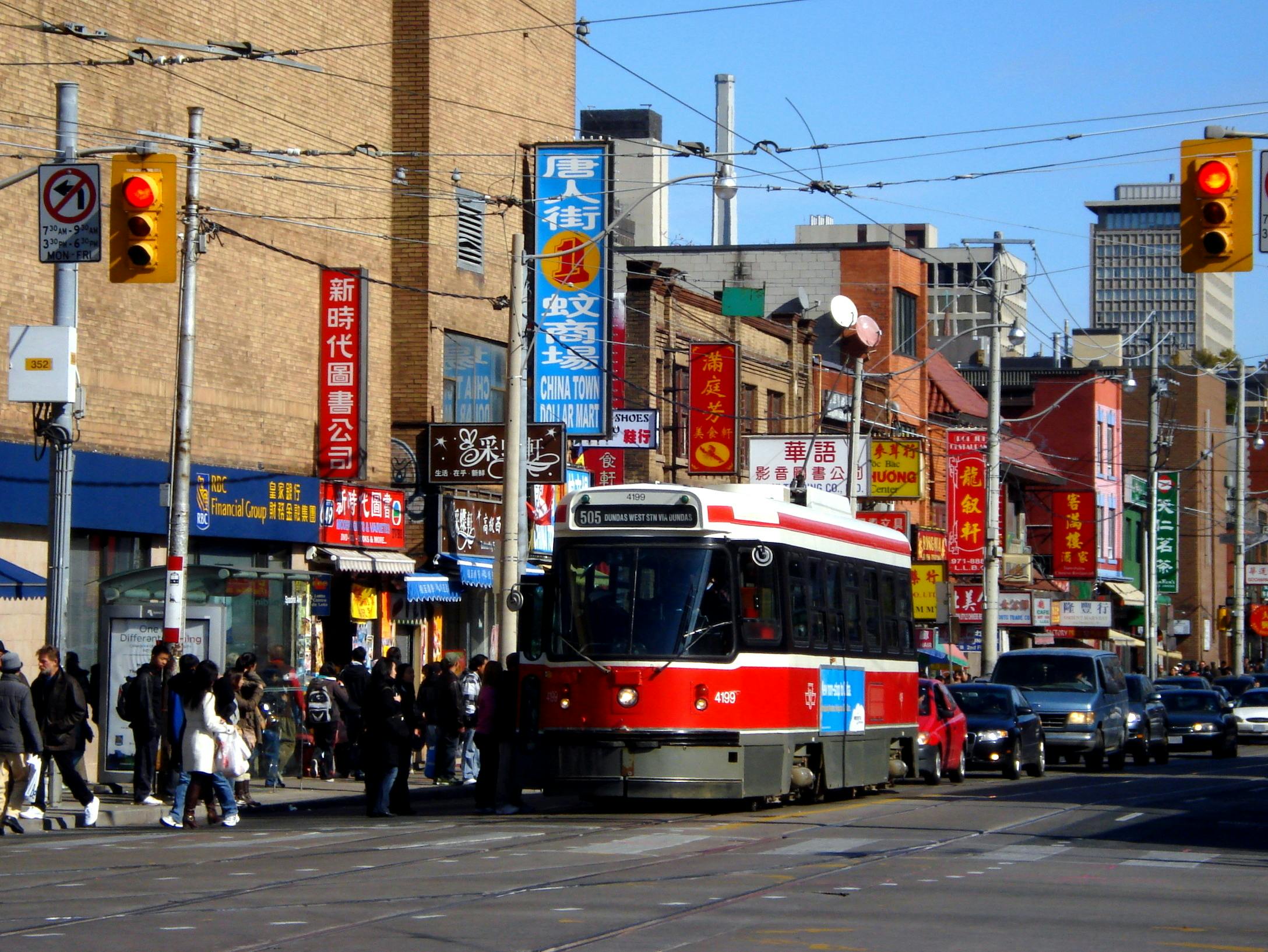 A red and white streetcar travels down a busy Chinatown street, surrounded by buildings with colorful signs and pedestrians.