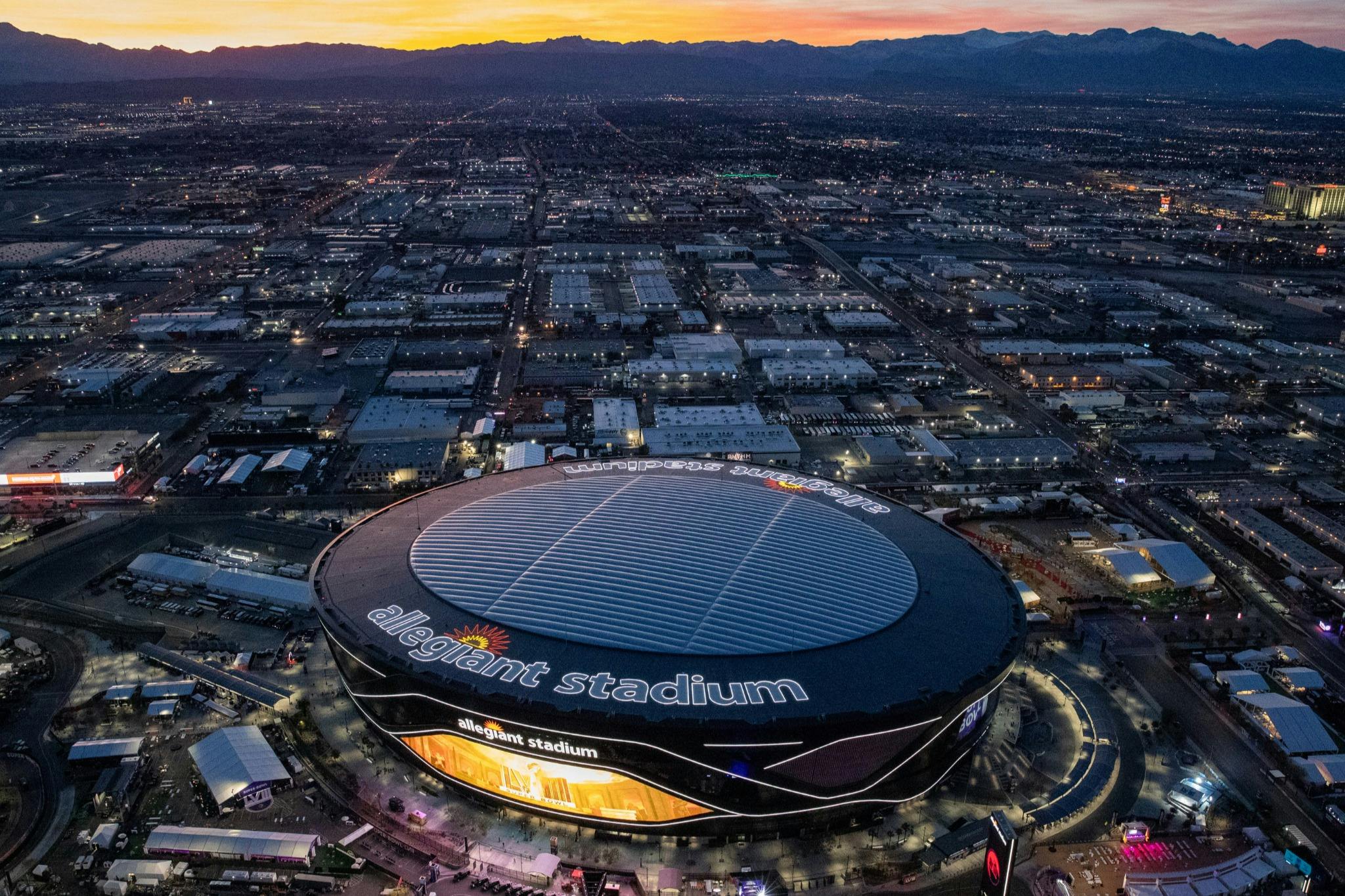 Aerial view of Allegiant Stadium illuminated at dusk, surrounded by city buildings and distant mountains under a colorful sky.
