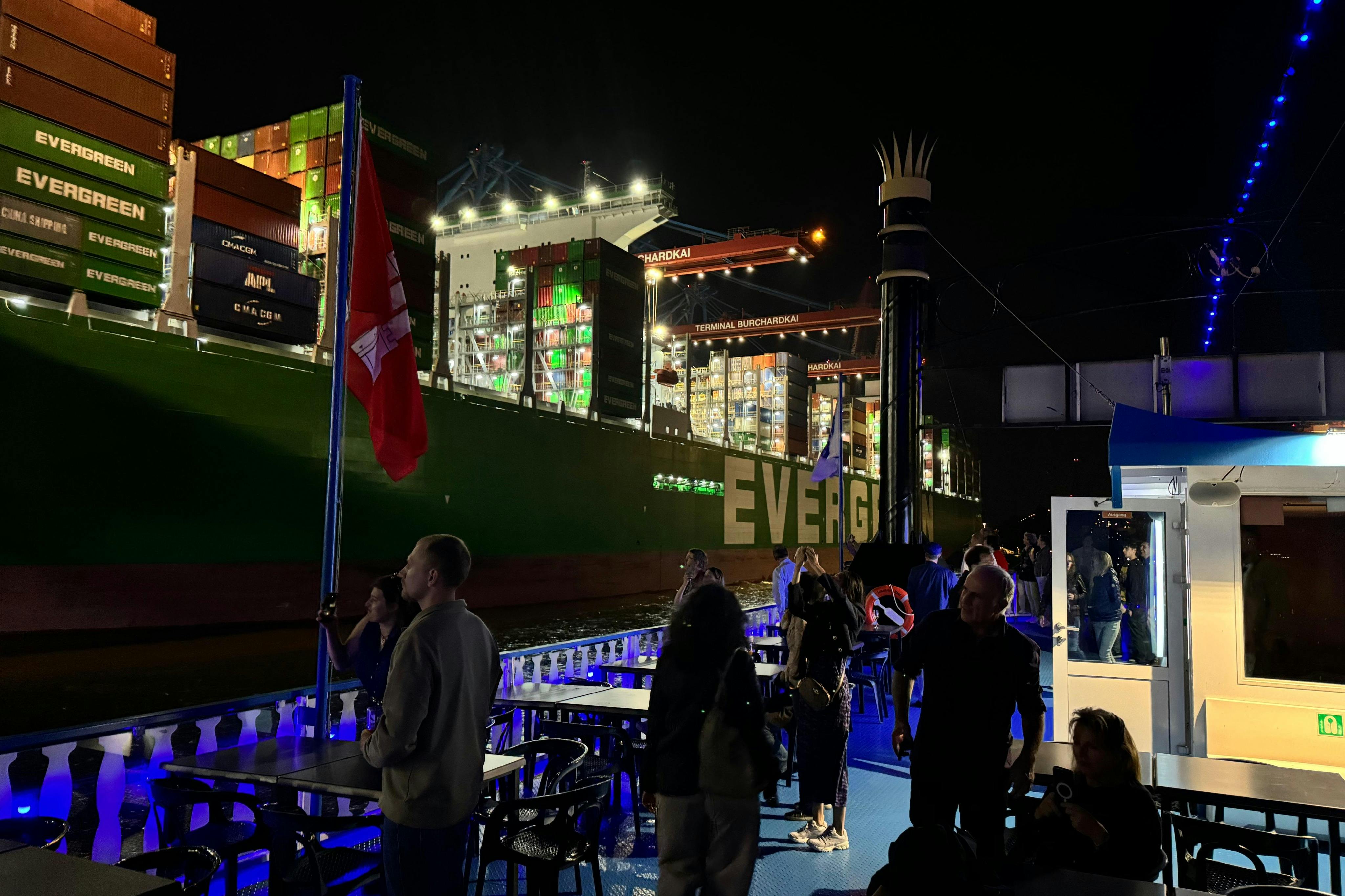 Ludzie stojący i siedzący nocą przy stołach na zewnętrznym pokładzie, z jasno oświetlonym statkiem towarowym w tle.