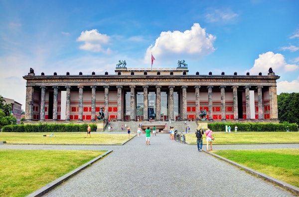 Large neoclassical building with columns and statues, people walking and cycling on a path, and a flag on the rooftop, under a blue sky.