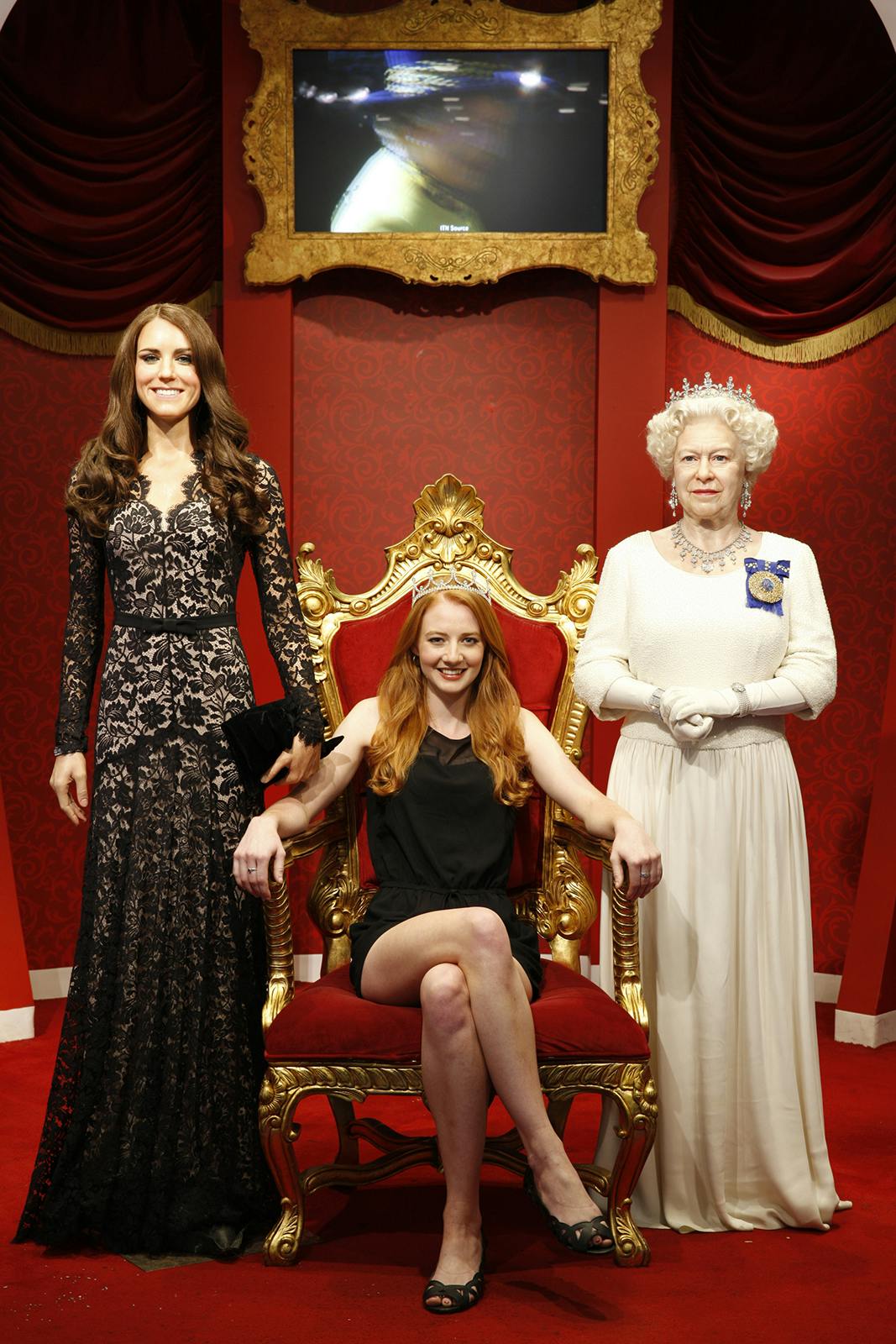 赤と金の背景を背景に、フォーマルな服装をした2人の女性の蝋人形の間に金の玉座に座っている女性。