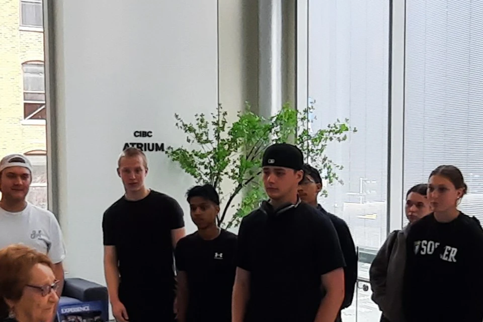 A group of people standing indoors near large windows, listening to a woman speaking. A poster is visible in the background.