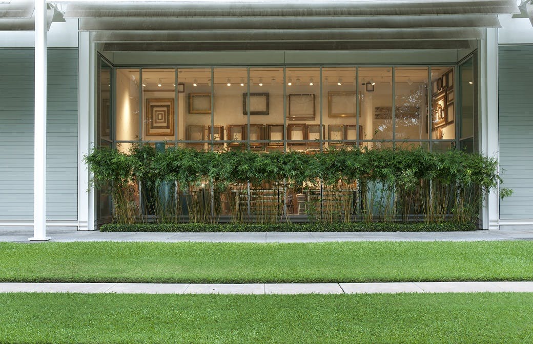 내부 벽에 액자 모양의 예술품이 있는 유리로 마감된 건물로, 높은 식물과 녹지가 줄지어 있는 야외에서 볼 수 있습니다.