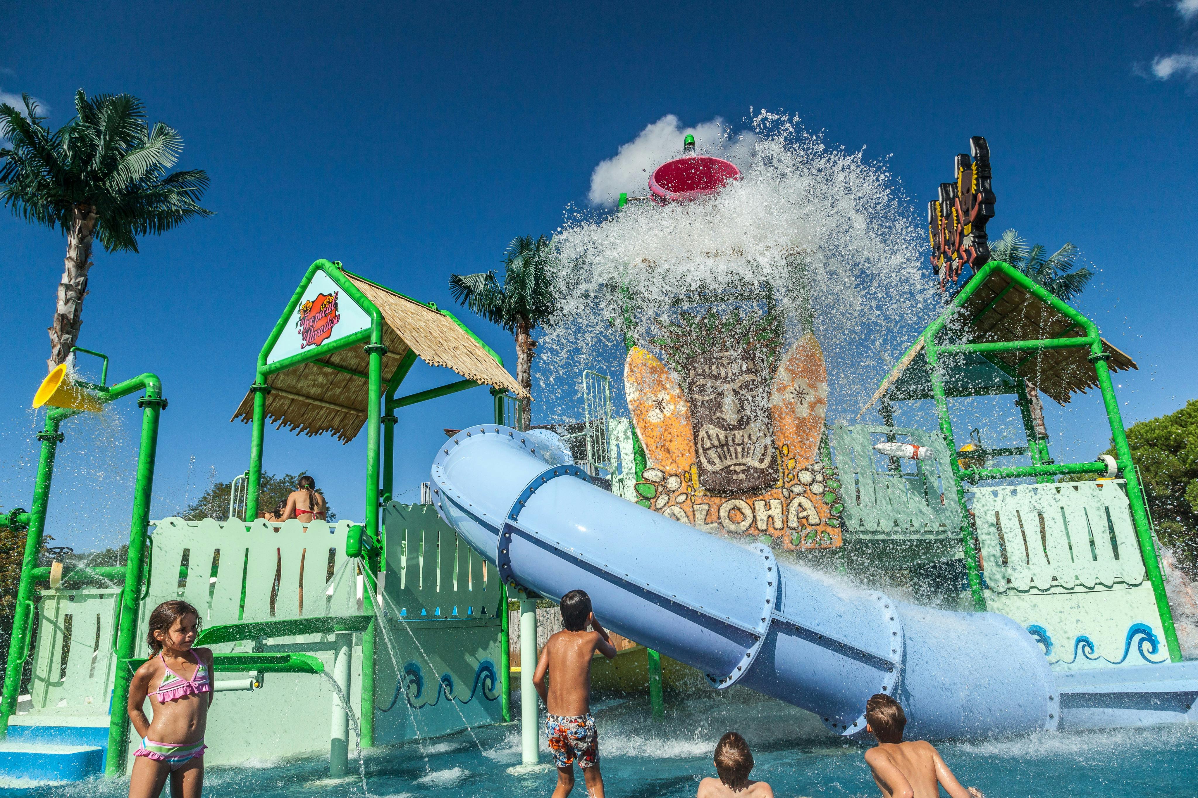 Des enfants jouant dans un parc aquatique avec des toboggans, des éclaboussures et un décor sur le thème du tiki sous un ciel bleu.