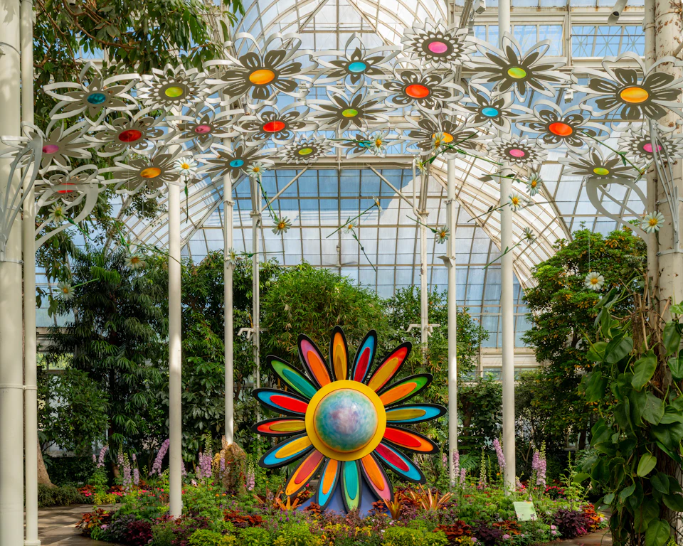 A colorful flower sculpture in a greenhouse surrounded by plants, with decorative flower shapes suspended overhead.