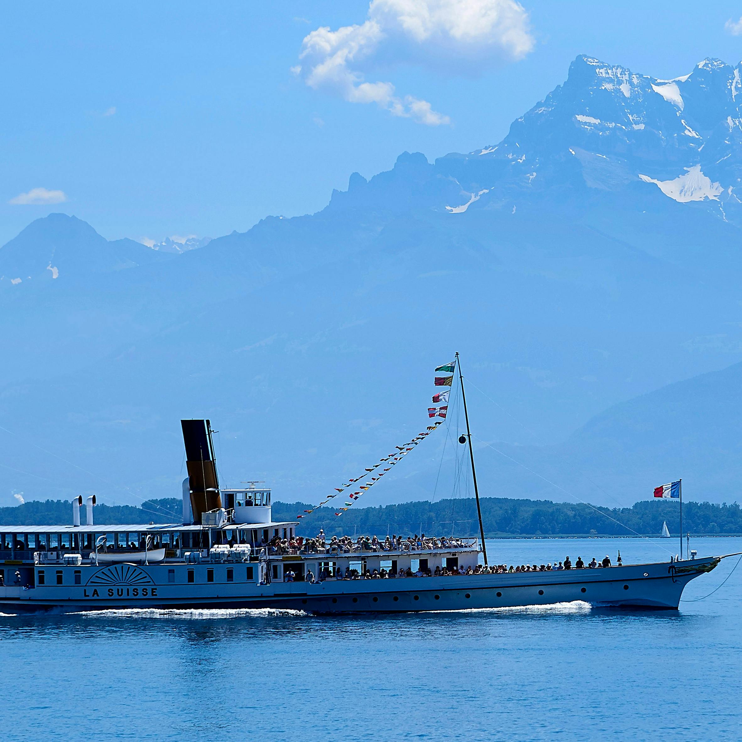 一艘名为“La Suisse”的船在平静的湖面上航行，背景是晴朗的蓝天下的群山。