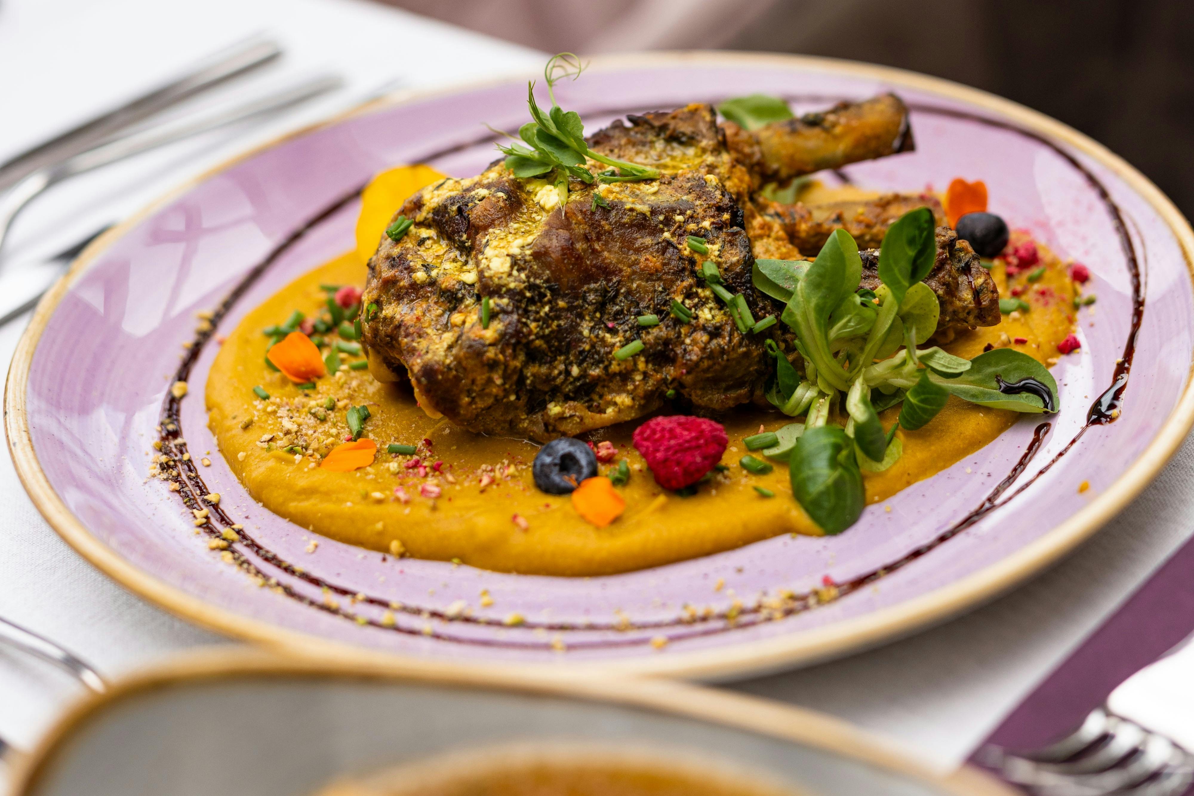 Un plat plat amb una picada de xai a la graella sobre un llit de puré daurat, guarnit amb verdures, baies i flors comestibles.