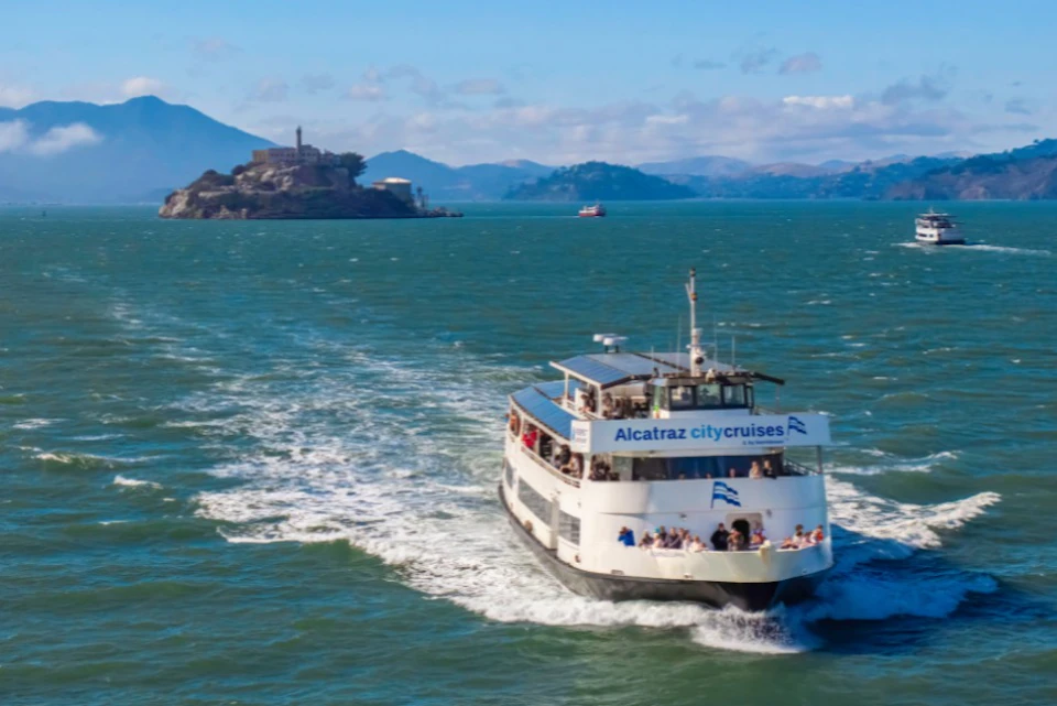 A ferry labeled "Alcatraz City Cruises" sails on water, with Alcatraz Island and a distant shoreline in the background.