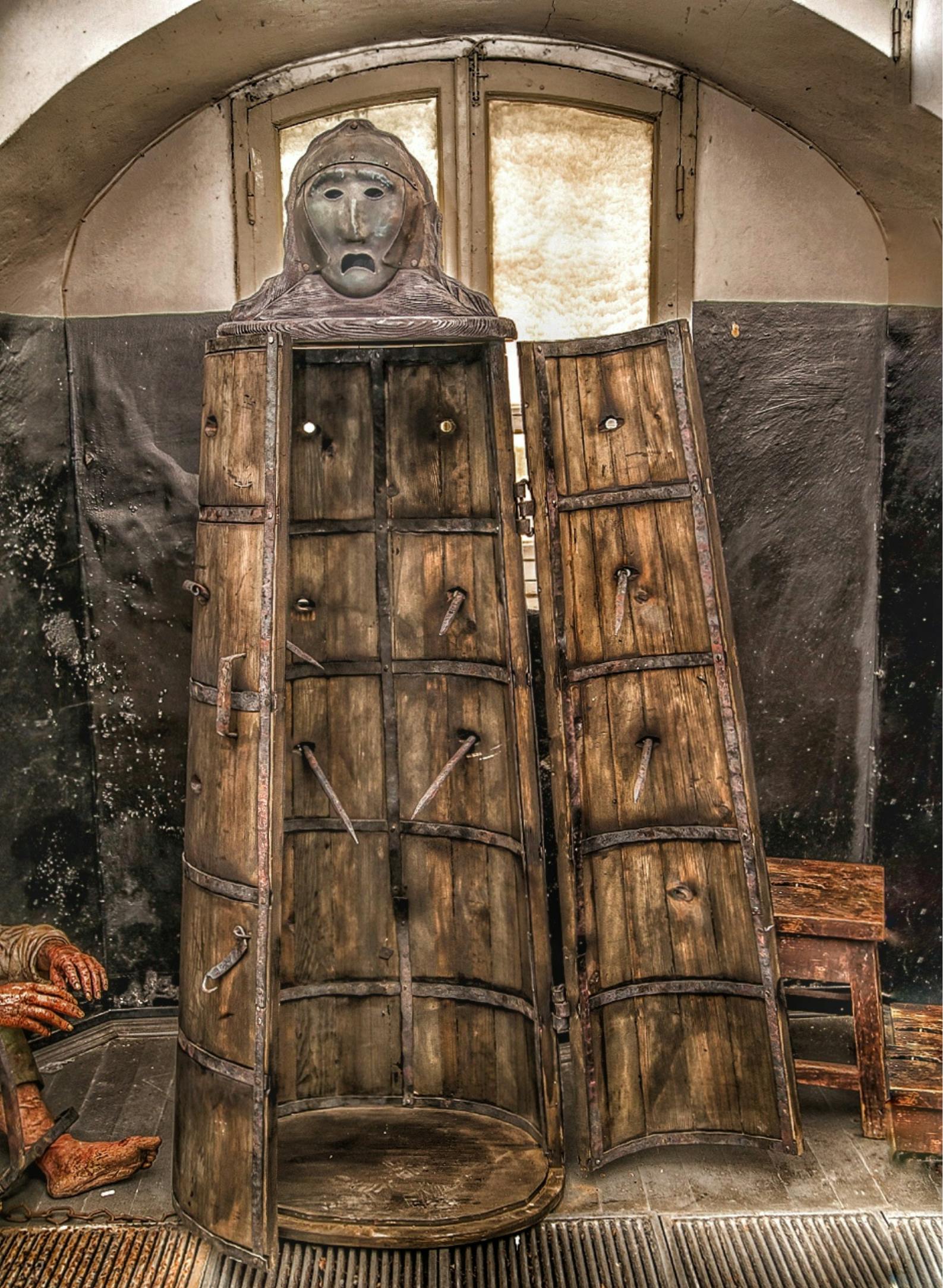 Otevři mučící zařízení Iron Maiden s lidskou tváří na vrchu, umístěné ve slabě osvětlené místnosti s kamennými zdmi a dřevěnou židlí poblíž.