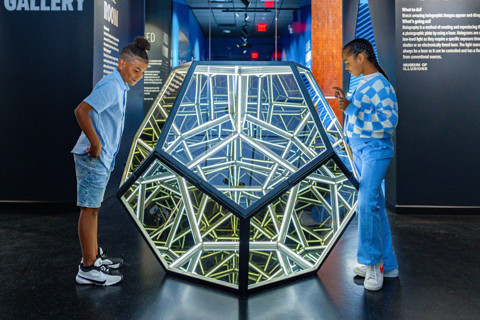 A child examines a geometric, illuminated sculpture with mirrored surfaces in a modern gallery space.