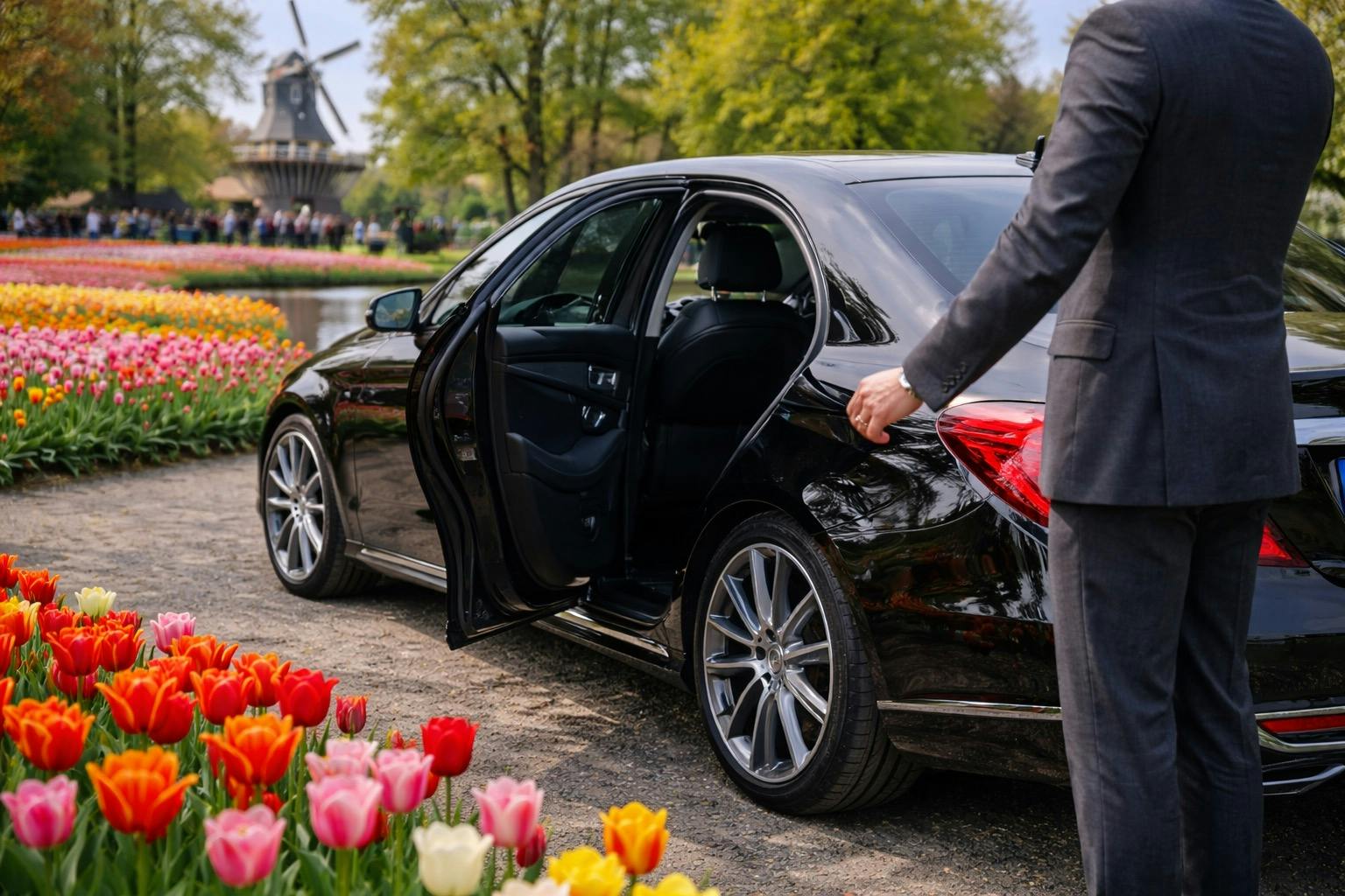 Czarny luksusowy samochód z otwartymi drzwiami, trzymającą go osobą w garniturze, w pobliżu tulipanów i malowniczym tłem z drzewami i wiatrakiem.