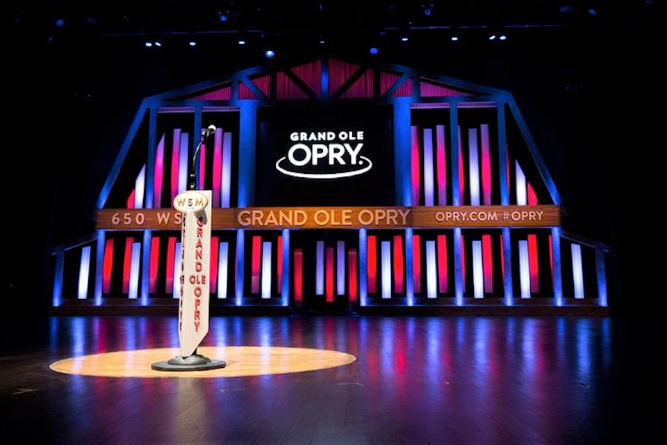 Grand Ole Opry stage with a vintage microphone stand under spotlights, surrounded by illuminated red and blue vertical lights.