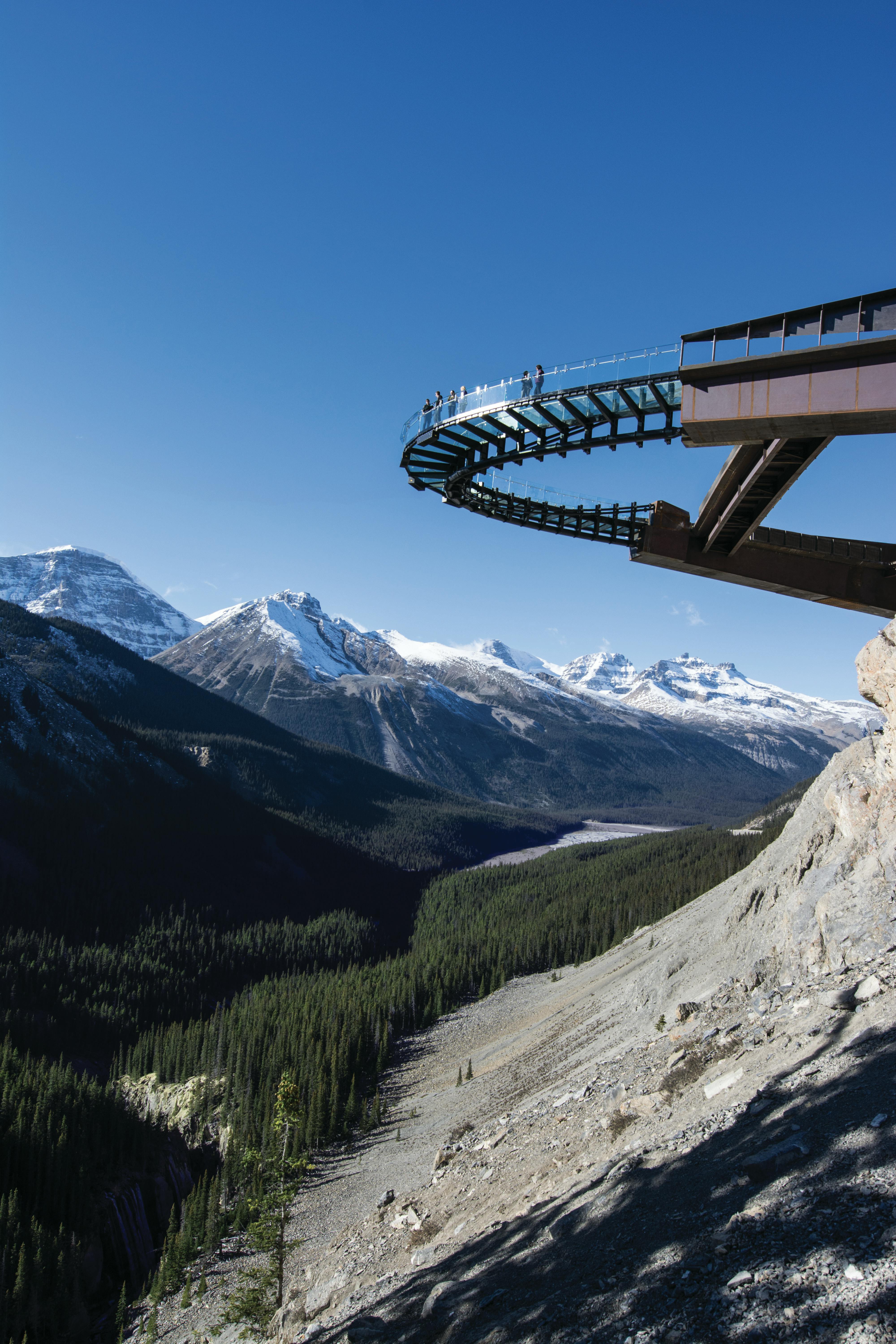 Glass-floored skywalk extending from a cliff, with people walking on it, set against a backdrop of snow-capped mountains and blue sky.