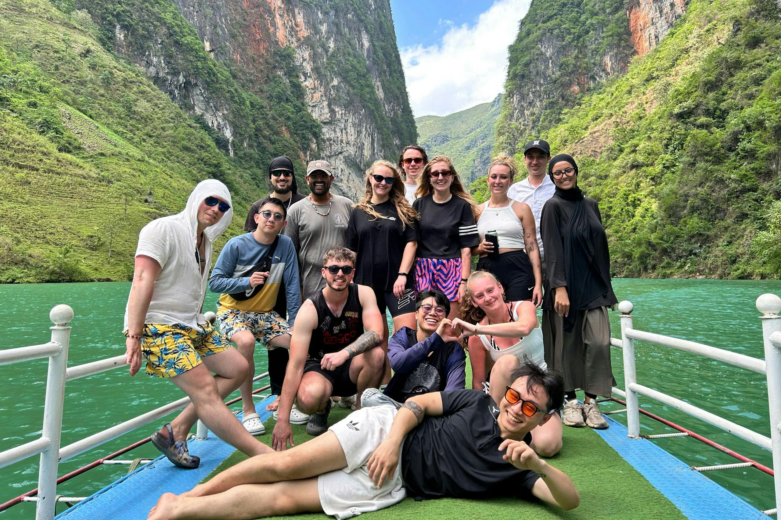 En gruppe mennesker, der smiler og poserer sammen på en båd, omgivet af et naturskønt, frodigt, bjergrigt landskab med en blå himmel.