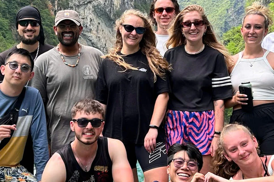 A group of people smiling and posing together on a boat, surrounded by a scenic, lush, mountainous landscape with a blue sky.