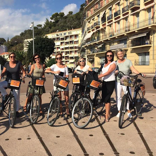 Sex kvinnor står med cyklar en solig dag, med färgglada byggnader och träd i bakgrunden.